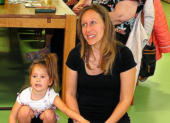 Eine Frau und ein junges Mädchen sitzen zusammen auf dem Boden. Die Frau lächelt und sieht zur Kamera, während das Mädchen geradeaus schaut. Hinter ihnen steht ein Holztisch und eine Person in einem Blumenhemd.