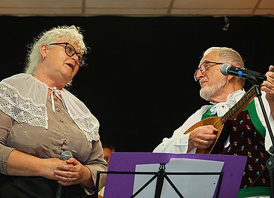 Eine ältere Frau mit weißen Haaren und Brille steht neben einem älteren Mann mit Schnurrbart, der eine Mandoline spielt. Sie halten beide Mikrofone. Dahinter steht ein Notenständer mit Musiknoten.
