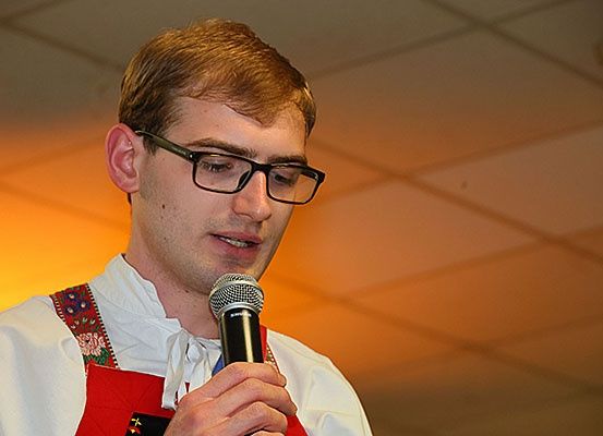 Ein Mann in einem weißen Hemd mit rotem Schürze und Brille hält ein Mikrofon und scheint zu sprechen.