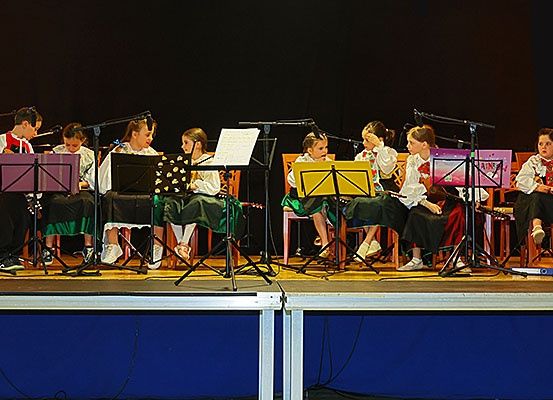 Eine Gruppe von Kindern spielt auf der Bühne. Sie spielen verschiedene Instrumente, darunter Flöten, Gitarren und Violinen. Die Kinder sind in passenden Uniformen gekleidet und sitzen auf Stühlen mit Notenständern vor ihnen. Die Bühne ist von Bühnenleuchten beleuchtet, und der Hintergrund ist dunkel.