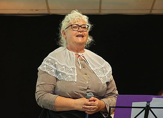 Eine Frau mit weißem Haar und Brille steht auf der Bühne, hält ein Mikrofon, lächelt und trägt einen Spitzenkragen. Hinter ihr ist ein Notenständer mit Noten zu sehen.