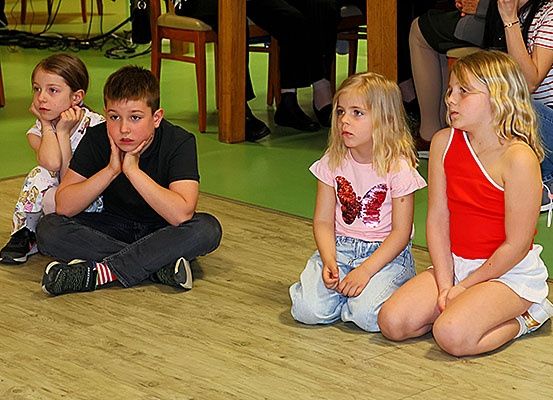 Vier Kinder, zwei Mädchen und zwei Jungen, sitzen auf einem Boden, vielleicht in einem Klassenzimmer, und scheinen etwas aufmerksam zu hören.