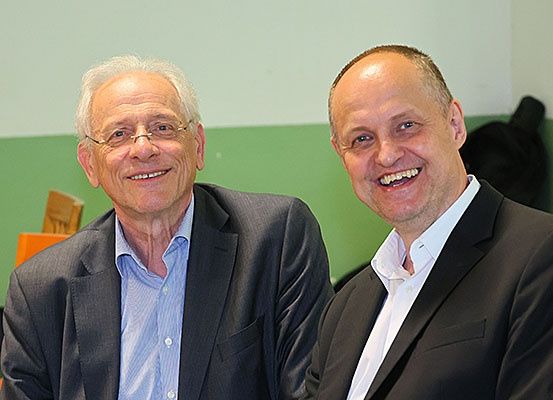 Zwei Männer sitzen und lächeln. Der Mann auf der linken Seite trägt eine Brille und ein hellblaues Hemd. Der Mann auf der rechten Seite trägt einen schwarzen Anzug.