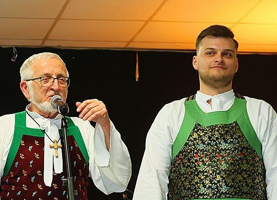 Zwei Männer stehen zusammen, einer mit einem Mikrofon, in traditioneller Kleidung. Der ältere Mann hält ein Mikrofon, während der jüngere Mann in die Kamera lächelt.
