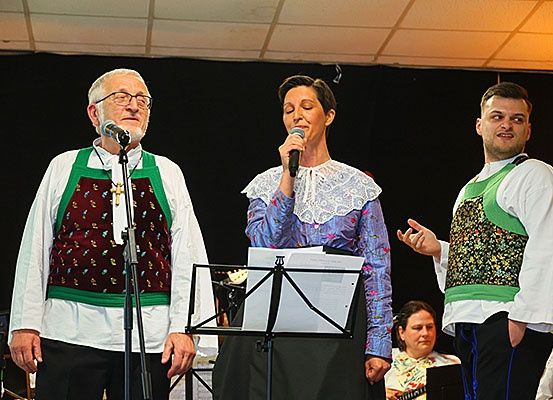 Drei Personen treten auf der Bühne auf. Der Mann links singt in ein Mikrofon. Die Frau in der Mitte hält ein Mikrofon und singt. Der Mann rechts schaut die Frau an.