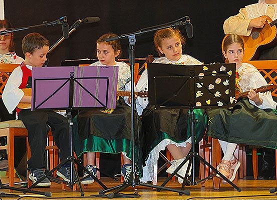 Kinder in traditioneller Kleidung spielen Gitarren und sitzen auf der Bühne unter einem Mikrofonständer.