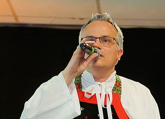 Ein Mann in einer weißen Robe und mit einer Brille singt in ein Mikrofon. Er trägt eine Schürze in Rot und Weiß.