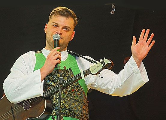 Ein Mann mit einer Gitarre tritt auf der Bühne auf, hält ein Mikrofon und hebt die Hand. Er ist in einer grünen Weste mit Blumenmuster gekleidet.