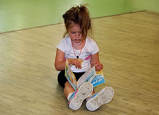 Ein junges Mädchen mit zerzaustem Haar sitzt auf dem Boden und ist in ein Buch vertieft. Sie trägt ein weißes T-Shirt und schwarze Shorts.