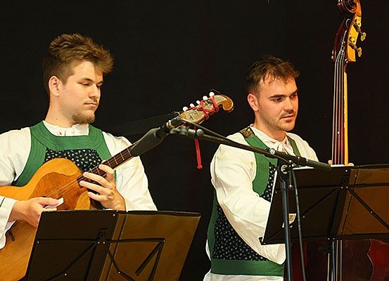 Zwei Männer in traditioneller Kleidung spielen Gitarre und Bass auf einer Bühne. Der Mann mit der Gitarre strummt und singt, während der andere den Bass hält und spielt.