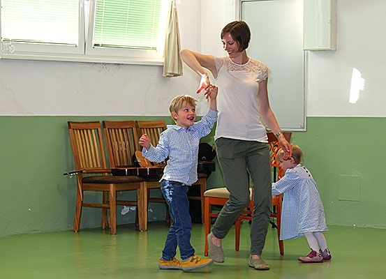 Eine Frau hält die Hände von zwei Kindern, möglicherweise in einem Klassenzimmer, mit Stühlen und einem Klavier im Hintergrund.