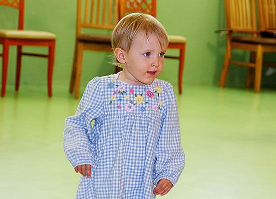 Ein kleines Kind in einem blau-weiß karierten Kleid steht in einem grünen Raum, mit Holzstühlen im Hintergrund.