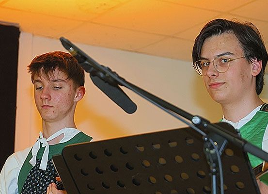 Zwei Jungen in grünen Schürzen und weißen Krawatten spielen Gitarren an einem Mikrofon auf der Bühne.