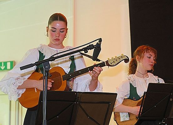 Zwei Frauen spielen Gitarren vor einem Mikrofonständer. Beide tragen weiße Blusen mit grünem Besatz. Die Frau auf der linken Seite schaut nach unten. Die Frau auf der rechten Seite schaut auf etwas rechts. Dahinter ist ein schwarzer Hintergrund.