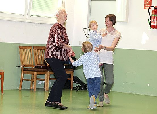Zwei Frauen und zwei Kinder tanzen in einem Raum mit grünen Wänden, Stühlen und einem Feuerlöscher.