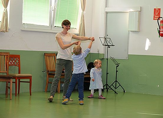 Eine Frau führt zwei Kinder in einem Tanz an, wobei ein Feuerlöscher und ein Notenständer im Hintergrund zu sehen sind.