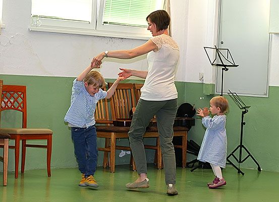 Eine Frau und zwei Kinder tanzen in einem Raum mit grünen Wänden, Stühlen und einem Notenständer. Die Frau hält die Hände der Kinder.