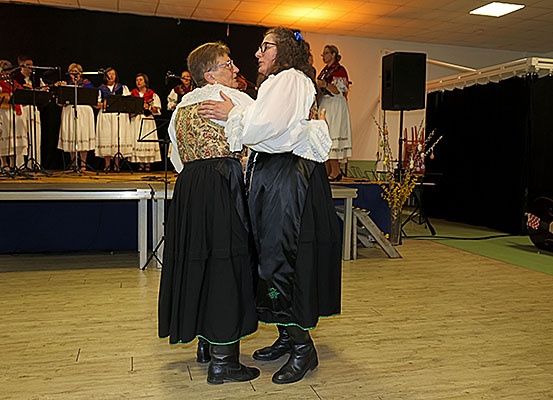 Zwei Frauen umarmen sich auf einer Tanzfläche während einer Aufführung, mit einem Chor auf der Bühne hinter ihnen.