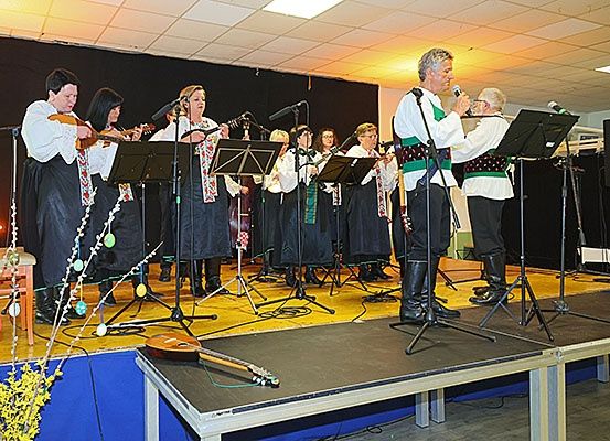 Eine Gruppe von Musikern tritt auf einer Bühne auf, einige spielen Instrumente, während andere singen. Die Bühne ist mit Musikinstrumenten und Blumen geschmückt.