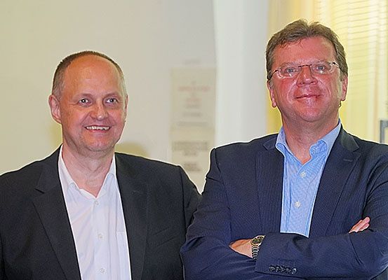 Zwei Männer stehen nebeneinander und tragen beide Anzüge. Der Mann auf der linken Seite lächelt und der Mann auf der rechten Seite hat die Arme verschränkt. Sie scheinen für ein Foto zu posieren.