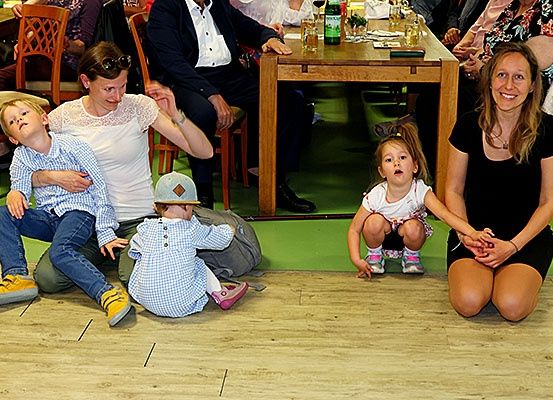 Eine Gruppe von Familien, einschließlich einer Frau mit zwei Kindern und einer anderen Frau mit einem kleinen Mädchen, sitzen auf dem Boden in einem Raum. Hinter ihnen steht ein langer Holztisch mit Essen und Getränken.