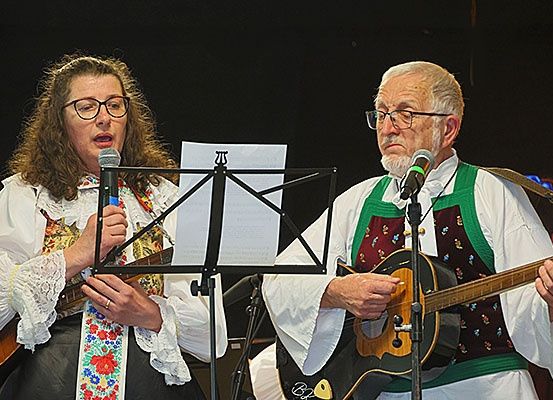 Zwei Musiker auf der Bühne, eine Frau singt in ein Mikrofon und ein Mann spielt Gitarre, mit Notenständern und Instrumenten vor ihnen.
