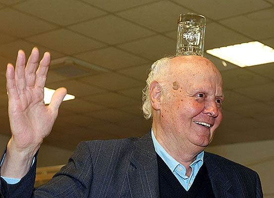 Ein älterer Mann mit einem Glas auf dem Kopf, lächelnd und winkend, gekleidet in einen Anzug und ein blaues Hemd.