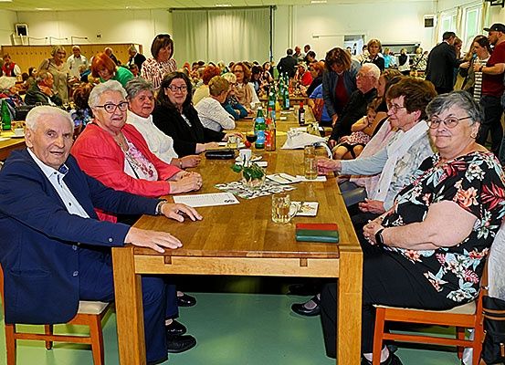 Eine Gruppe älterer Menschen sitzt an einem langen Tisch in einem Saal. Sie haben Getränke und einige Papiere auf dem Tisch.