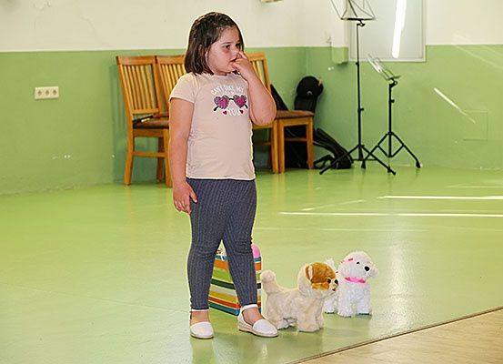 Ein Mädchen steht in einem Raum mit grünen Wänden und beißt sich auf die Nägel. Sie trägt ein rosa T-Shirt und graue Leggings. Zwei Spielzeughunde liegen auf dem Boden. Hinter ihr sind Holzstühle und ein Notenständer zu sehen.
