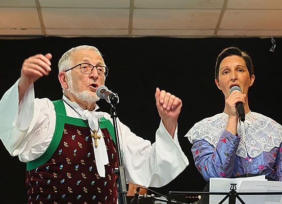 Zwei Personen, ein Mann und eine Frau, stehen auf der Bühne und singen. Der Mann trägt ein weißes Hemd mit einer rot-grünen Weste. Die Frau ist in einem blauen Hemd mit einem weißen Spitzenkragen gekleidet. Beide halten Mikrofone. Hinter ihnen sind Musikinstrumente zu sehen.
