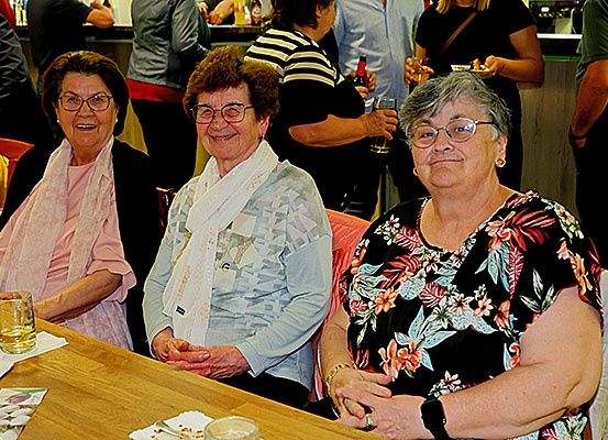 Drei ältere Frauen mit Brille und Lächeln sitzen an einem Tisch in einem Restaurant. Sie haben Getränke vor sich und es stehen und sitzen Leute hinter ihnen.