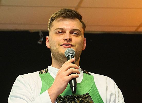 Ein Mann mit kurzen Haaren, der ein weißes Hemd und grüne Hosenträger trägt, hält ein Mikrofon in der rechten Hand und scheint auf der Bühne zu spielen.