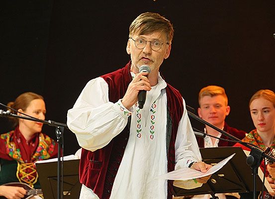 Ein Mann spricht in ein Mikrofon, während er vor einer Gruppe von Musikern in traditioneller Kleidung steht.