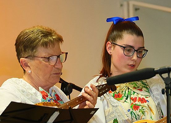 Zwei Frauen in traditionellen Kostümen spielen Gitarren und singen in Mikrofone auf einer Bühne.