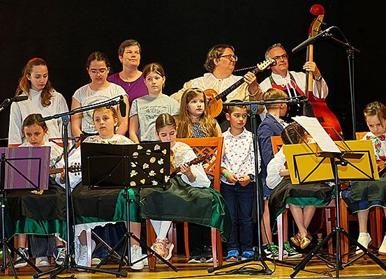 Eine Gruppe von Kindern und Erwachsenen auf einer Bühne spielt Gitarren und andere Instrumente. Die Kinder sitzen auf Stühlen mit Notenständern vor ihnen. Zwei Erwachsene stehen hinter den Kindern, einer spielt Cello und der andere Gitarre.