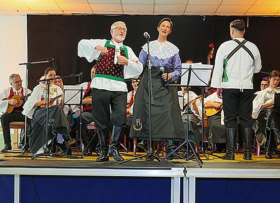 Eine Gruppe von Musikern tritt auf einer Bühne auf. Einer singt in ein Mikrofon. Ein anderer spielt Gitarre. Dahinter sitzt ein Cellospieler auf einem Stuhl. Andere Musiker sitzen um sie herum.