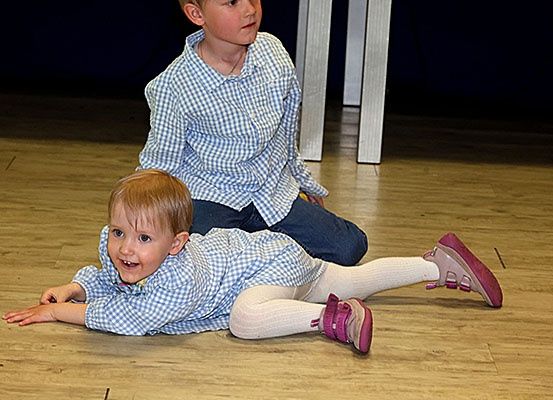 Zwei Kinder sitzen auf dem Boden. Der Junge rechts schaut den anderen Jungen an. Der Junge links liegt mit erhobenen Beinen auf dem Boden.