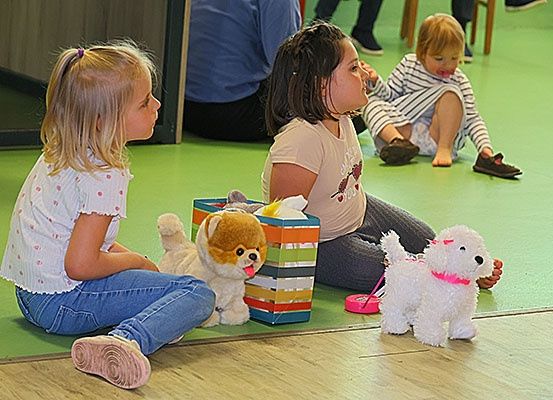 Drei junge Mädchen sitzen auf einem grünen Boden, zwei mit Plüschhunden. Ein Mädchen schaut auf ihren Hund, ein anderes schaut auf eine Spielzeugkiste und das dritte Mädchen hat ihre Zunge rausgestreckt.