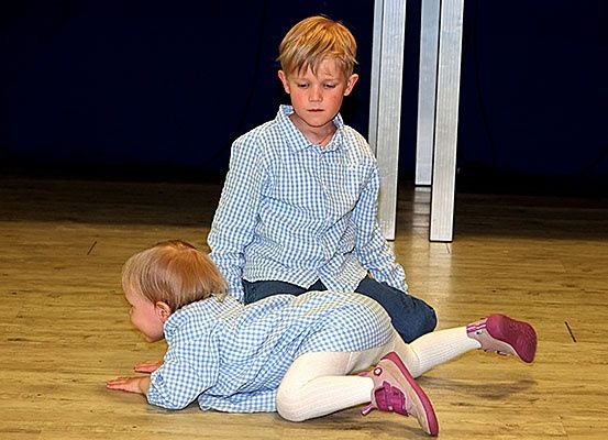 Zwei Kinder sind auf einem Holzboden, eines sitzt mit gekreuzten Beinen und das andere liegt auf dem Rücken. Beide tragen karierte Hemden und Jeans, wobei das sitzende Kind rosa Schuhe trägt.