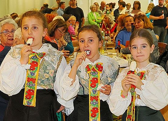 Drei Kinder in traditioneller Kleidung essen Eis bei einer Zusammenkunft mit anderen Menschen hinter ihnen.