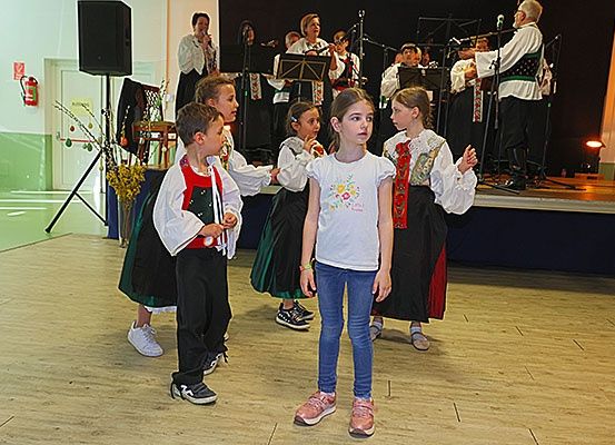 Eine Gruppe von Kindern in traditioneller Kleidung führt einen Tanz auf der Bühne auf, während eine Band im Hintergrund spielt.