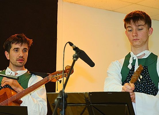 Zwei Männer spielen in einem Raum Musikinstrumente. Ein Mann hält eine Gitarre, während der andere eine Geige spielt. Sie stehen vor einem Mikrofon auf einem Ständer. Sie tragen traditionelle Kleidung. Der Raum hat eine weiße Wand und einen schwarzen Hintergrund.