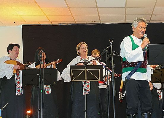 Eine Gruppe Musiker tritt auf der Bühne mit traditioneller Kleidung auf, mit einem singenden Mann und zwei Frauen, die Gitarre spielen, vor einem schwarzen Hintergrund.
