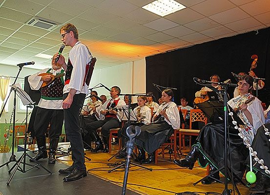 Eine Band spielt auf der Bühne mit verschiedenen Instrumenten; der Leadsänger steht an einem Mikrofon, während andere Gitarren, Celli und Violinen spielen. Das Publikum sitzt auf Stühlen, und es gibt Notenständer und Mikrofone auf der Bühne.