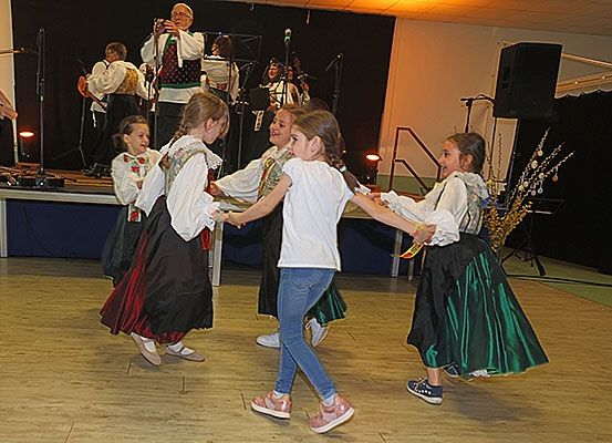 Eine Gruppe junger Mädchen tanzt auf einem Holzboden, möglicherweise bei einer kulturellen Veranstaltung, während eine Band auf der Bühne hinter ihnen spielt.