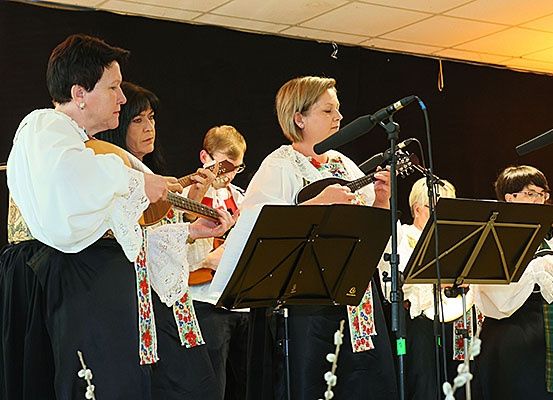 Eine Gruppe von Frauen in traditioneller Kleidung spielt Musikinstrumente, darunter Gitarren und Mandolinen, vor einem schwarzen Hintergrund. Sie stehen vor Mikrofonen und Notenständern.