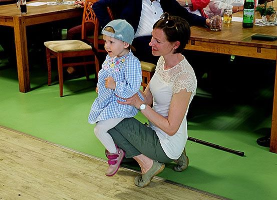 Eine Frau kniet mit einem Kind, einem Mädchen in einem blauen Kleid, auf ihrem Schoß. Hinter ihnen steht ein Tisch mit Gläsern und Flaschen.