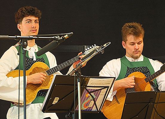 Zwei Männer in traditioneller Kleidung spielen Gitarren auf der Bühne. Beide haben Mikrofone vor sich. Dahinter befinden sich Notenständer mit Notenblättern. Der Hintergrund ist dunkel.