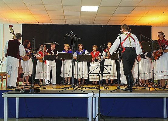 Eine Gruppe von Musikern, darunter ein Mann und mehrere Frauen, tritt auf einer Bühne auf. Sie halten Gitarren und stehen hinter einem Notenständer.