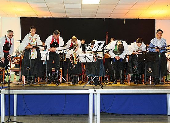Eine Gruppe von Musikern in traditioneller Kleidung tritt auf der Bühne auf und hält verschiedene Instrumente. Sie stehen vor einer Hintergrundkulisse mit einer Hintergrundbespannung.